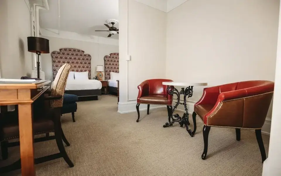 A hotel room with two beds, a wooden desk with a chair, a table with two red leather armchairs, beige carpet, and cream-colored walls. A ceiling fan and patterned headboards are visible in the background.