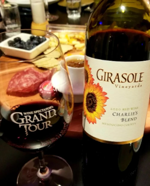 A bottle of Girasole Vineyards 2020 Charlie’s Blend red wine stands next to a wine glass and a charcuterie board—perfect for sharing cheese, crackers, blueberries, salami, and dip during a relaxing spring break gathering.