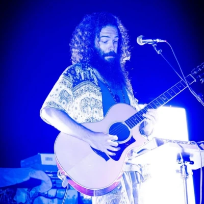 A man with long curly hair and a beard plays an acoustic guitar and sings into a microphone on stage, illuminated by blue lighting.