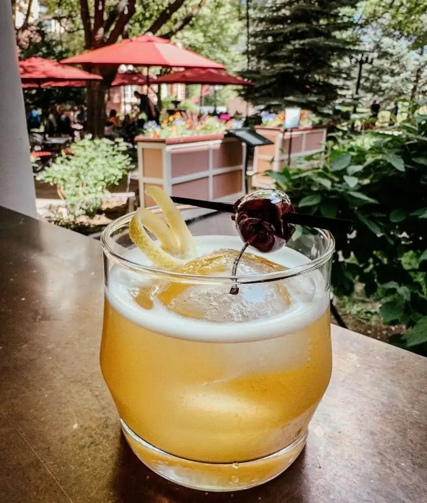 A yellow cocktail garnished with a lemon twist and a cherry sits on a table outdoors at the Coppertop Bar, surrounded by greenery, colorful flowers, and people relaxing beneath red umbrellas in the background.