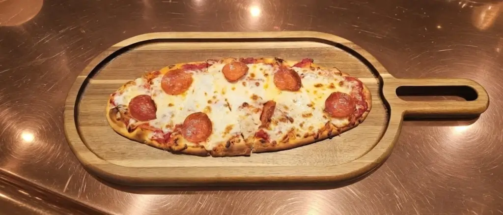 An oval-shaped pepperoni pizza with melted cheese is served on a wooden paddle board, resting atop the gleaming surface of the Coppertop Bar at Hotel Colorado.