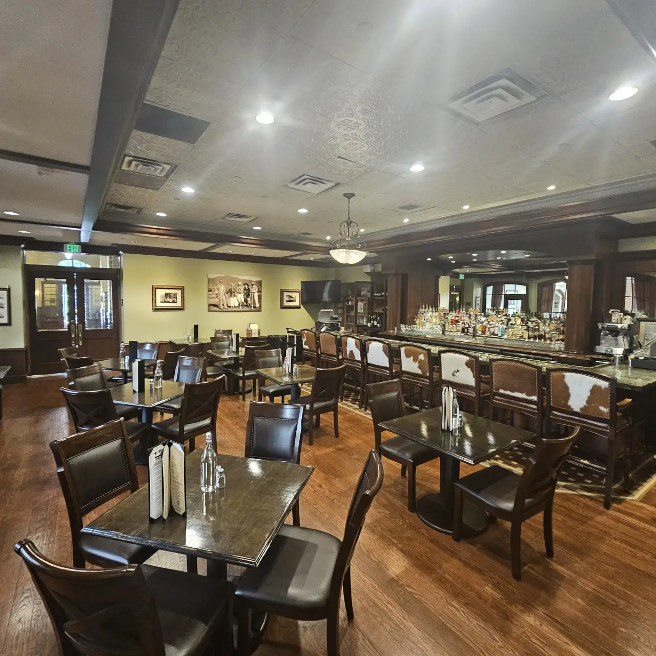 A spacious, well-lit restaurant dining area with dark wooden tables and chairs, a long bar counter, menus on tables, hardwood floors, and framed photos on cream-colored walls evoke the inviting ambiance of a classic polo lounge.