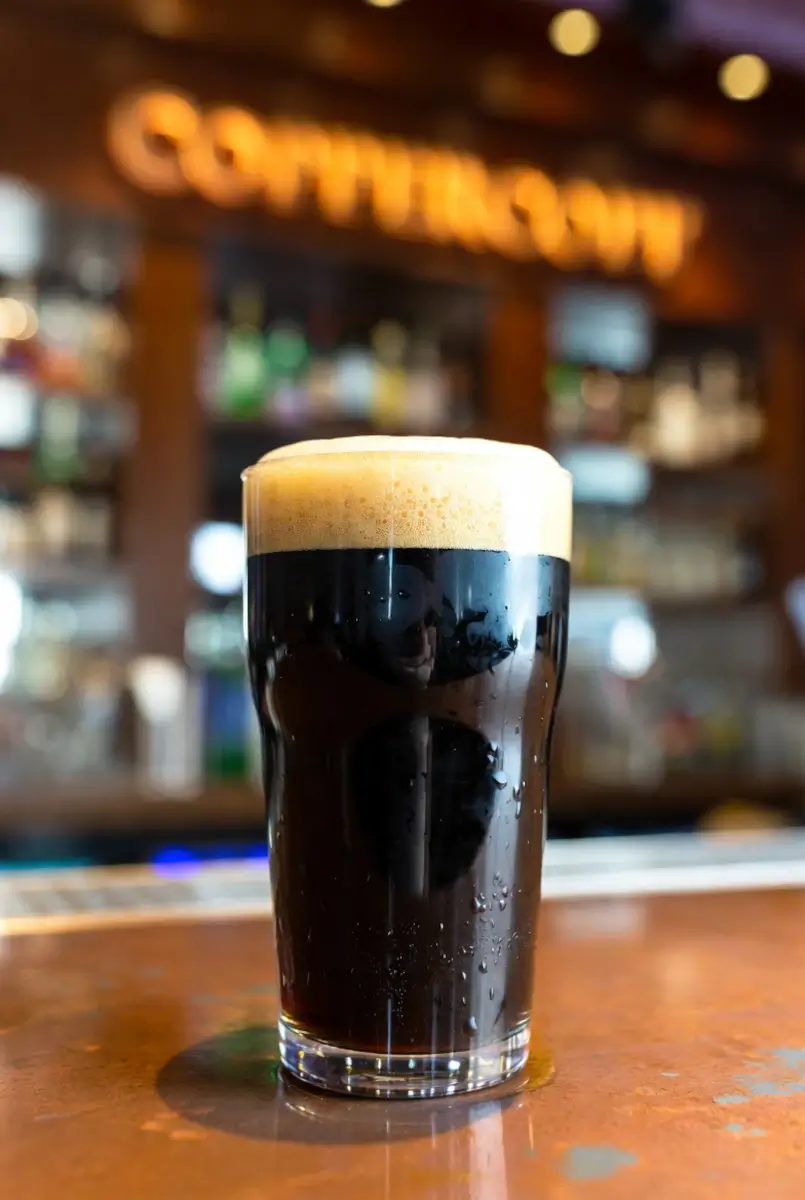 A pint glass filled with dark stout beer topped with a thick, creamy foam sits on a bar counter, with blurred bottles and a sign in the background.