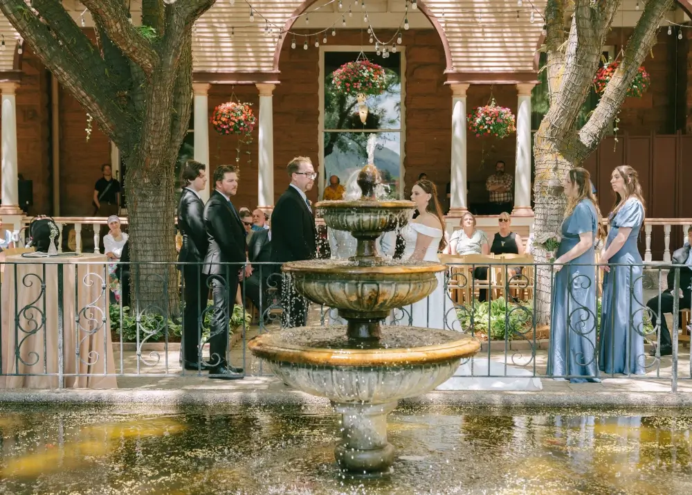 A wedding ceremony outdoors features a bride and groom facing each other near a tiered fountain, with bridesmaids, groomsmen, and guests seated in the background under string lights, creating a magical atmosphere that perfectly captures the romance of weddings.