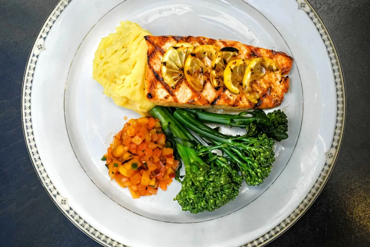 Grilled salmon topped with lemon slices, served with mashed potatoes, sautéed diced carrots, and broccolini on a white plate with a decorative rim.