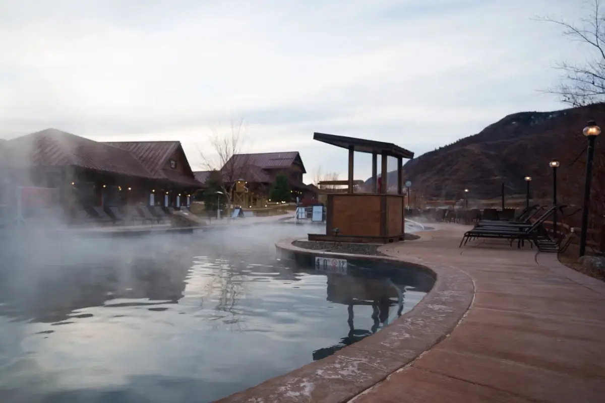 A steaming outdoor hot spring pool with lounge chairs along a curved walkway, surrounded by rustic buildings and the majestic peaks of Iron Mountain under a cloudy sky.