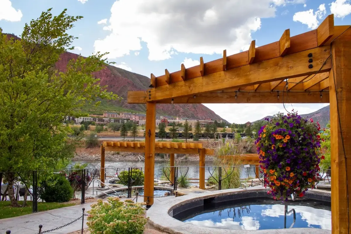 A hot tub sits under a wooden pergola near a river, surrounded by flowers and greenery, with Iron Mountain and clouds visible in the background.