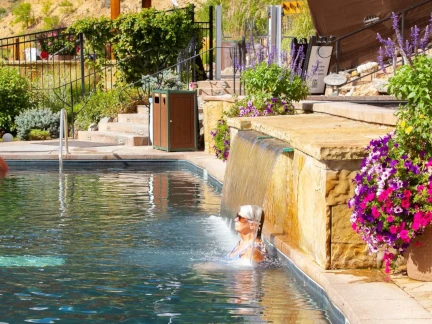 A person in sunglasses relaxes in a pool under a small waterfall at Iron Mountain, surrounded by colorful flowers and greenery on a sunny day.