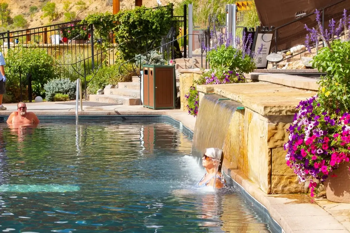 A person relaxes under a small waterfall at the edge of an outdoor pool, surrounded by flowers and stone walls reminiscent of Iron Mountain’s natural beauty, while another person swims nearby. Steps and greenery are visible in the background.