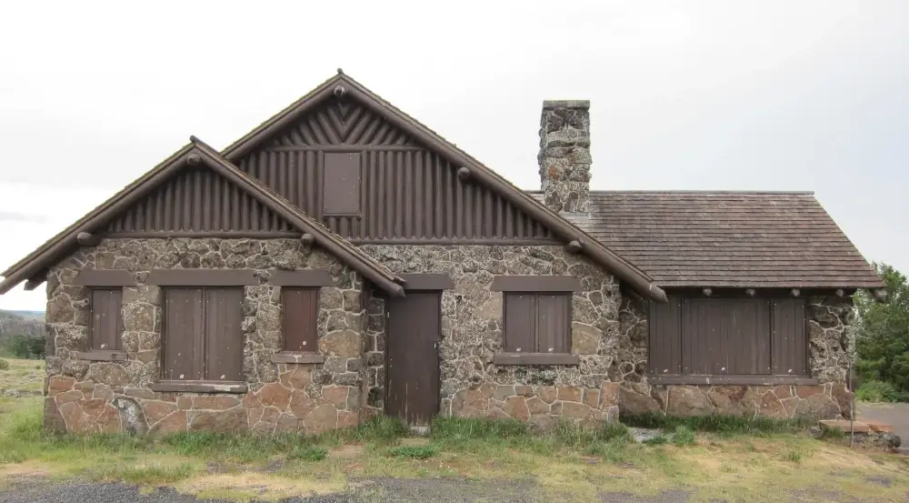 A stone cabin with boarded-up windows and doors, wooden gables, a central chimney, and a shingled roof, set on a grassy patch with cloudy skies in the background.