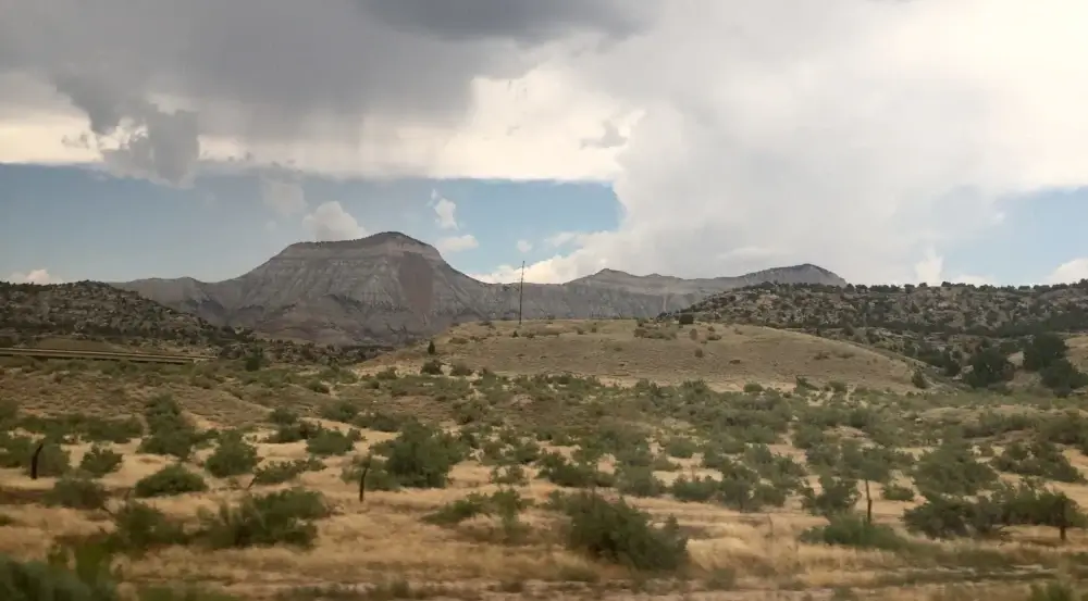 A dry, grassy landscape with scattered bushes, distant flat-topped mountains, and a cloudy sky overhead.