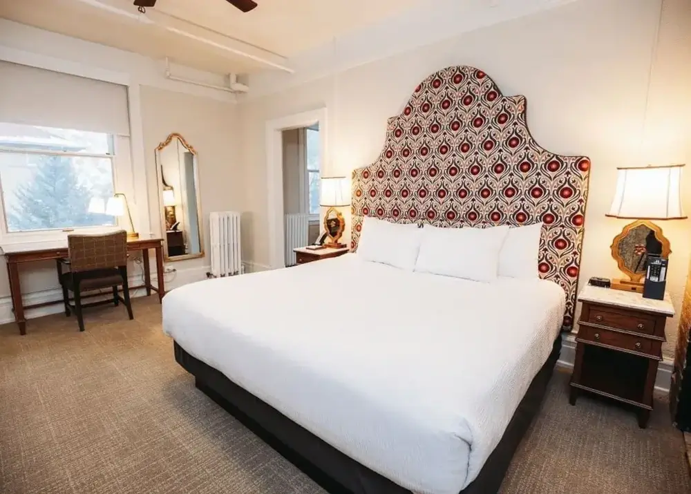 A hotel room with a large bed, patterned headboard, two nightstands with lamps, a desk and chair by the window, and a mirror on the wall. The decor is neutral with warm lighting, creating an inviting retreat perfect for relaxing after weddings or special celebrations.