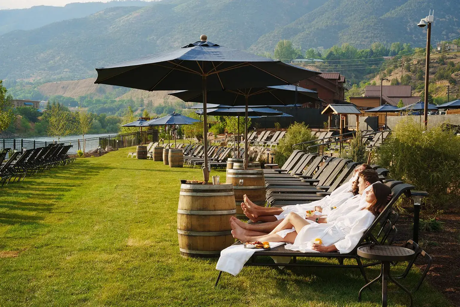Three people in white robes relax on lounge chairs under a large black umbrella on a grassy lawn, with the majestic Iron Mountain, a river, and more lounge chairs in the background. Food and drinks rest on a barrel nearby.