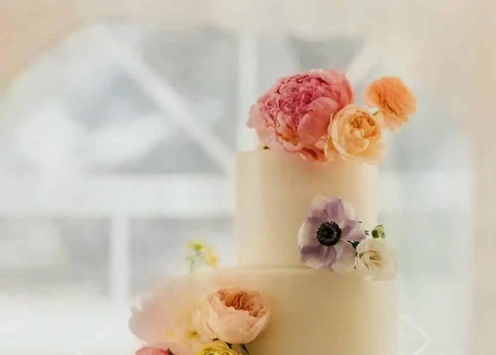 A two-tiered white wedding cake decorated with pastel-colored flowers, including pink, peach, and lavender blooms, set against a softly blurred background.