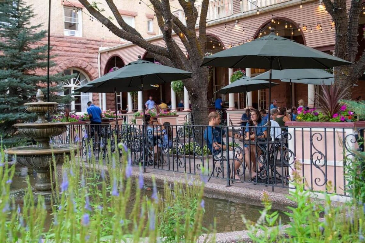 People sit at outdoor tables under umbrellas in a charming courtyard with a fountain, a locals' choice for relaxation. Surrounded by greenery and string lights, the setting exudes a relaxed atmosphere. The backdrop features a historic brick building with arched windows.